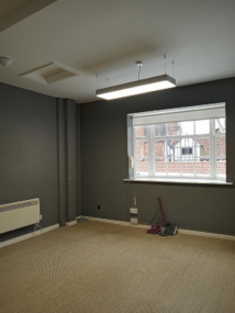 A photo in the office space, with carpeted floor, overhead lighting and a radiator, a window at the far end which looks to the street below