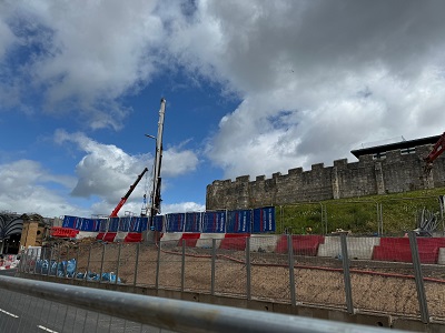 a view from the road of a piling machine on a building site. In the background, the City Walls sit upon a grassy hill, while in the foreground are acoustic barriers to dampen some of the noise